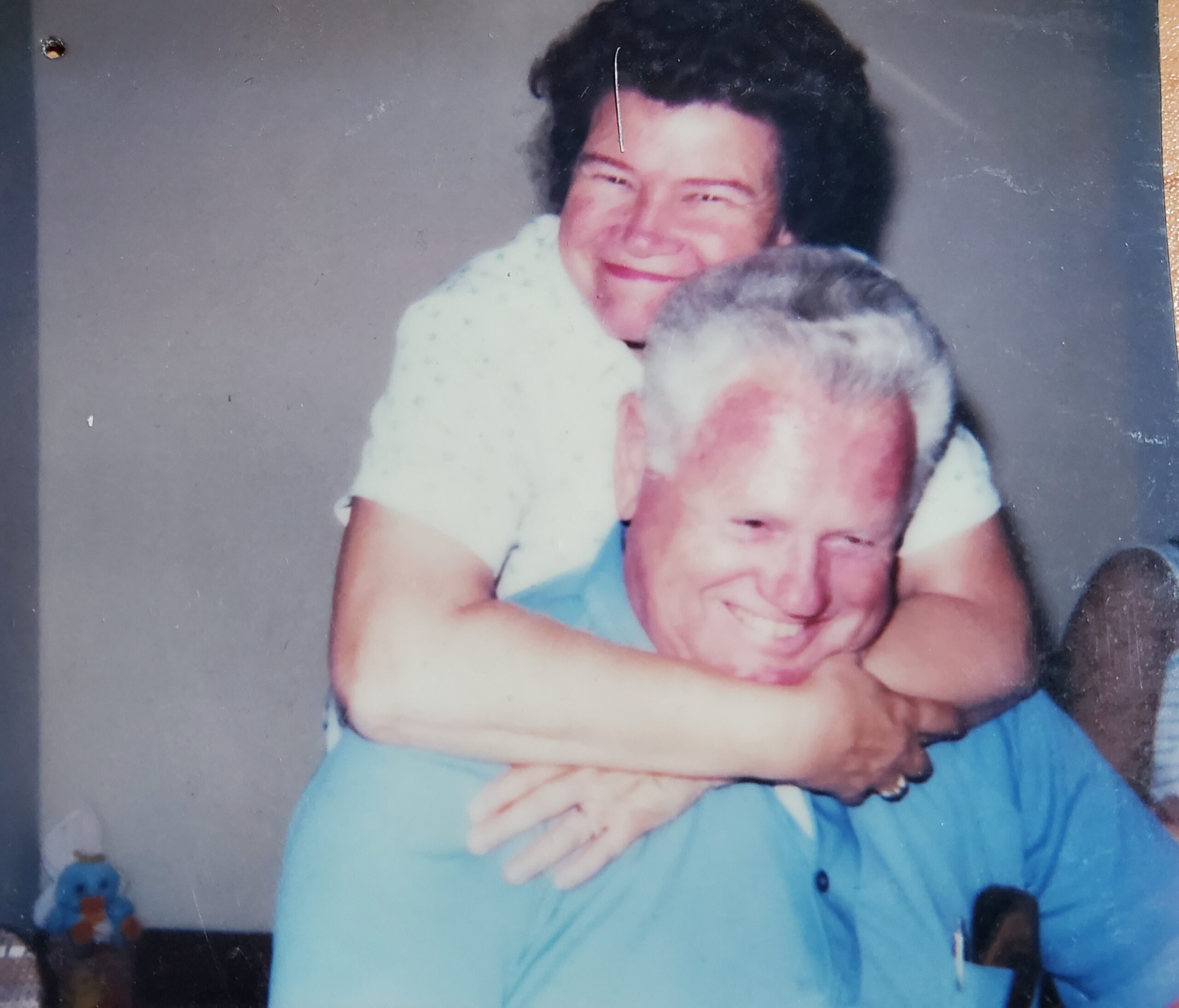 An older woman smiles and hugs an older man from behind. Both are seated indoors and appear happy.